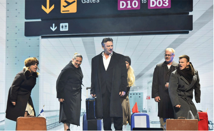 Anders stående i mörk rock och vit skjorta på flygplats, omgiven av fem resenärer. Alla sjunger.