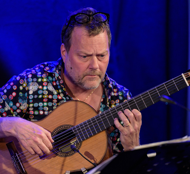 Bengt Magnusson i mönstrad skjorta spelar gitarr. Bakgrunden är blå och han har ett notställ framför sig.