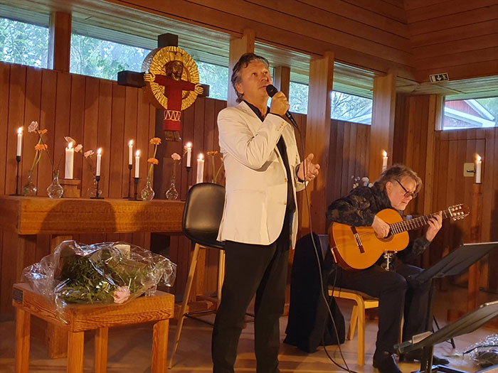 Anders Ekborg i vit kavaj sjunger, stående. Intill honom sitter Bengt Magnusson, klädd i svart, och spelar gitarr. I bakgrunden träinredning och tända ljus.